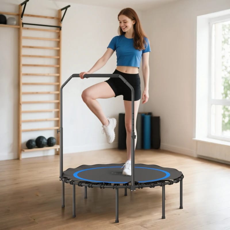 A woman exercises on a mini trampoline with a handrail, wearing a blue top and black shorts, in a bright fitness room with wooden flooring and gym equipment.