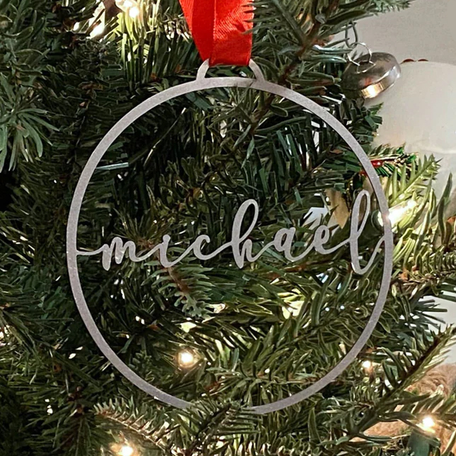 A circular Christmas tree ornament with the name \“Michael\“ inscribed in cursive, hanging from a red ribbon on a decorated evergreen branch with lights.