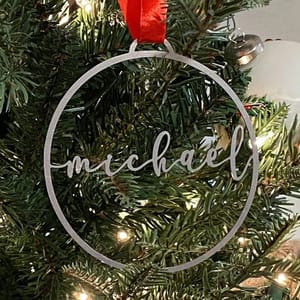 A circular Christmas tree ornament with the name \“Michael\“ inscribed in cursive, hanging from a red ribbon on a decorated evergreen branch with lights.
