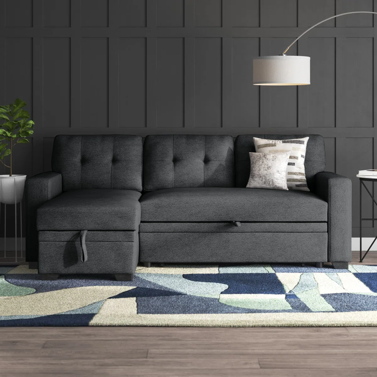 Dark gray sectional sofa with tufted cushioning, accompanied by a geometric rug and a floor lamp with a round shade. Nearby is a small side table with books and a potted plant.
