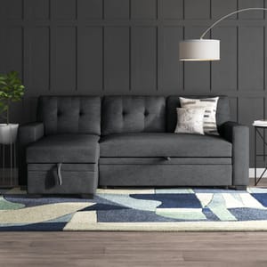 Dark gray sectional sofa with tufted cushioning, accompanied by a geometric rug and a floor lamp with a round shade. Nearby is a small side table with books and a potted plant.