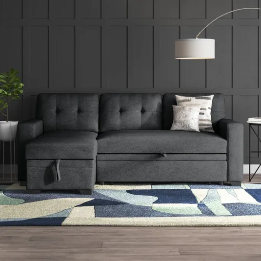 Dark gray sectional sofa with tufted cushioning, accompanied by a geometric rug and a floor lamp with a round shade. Nearby is a small side table with books and a potted plant.