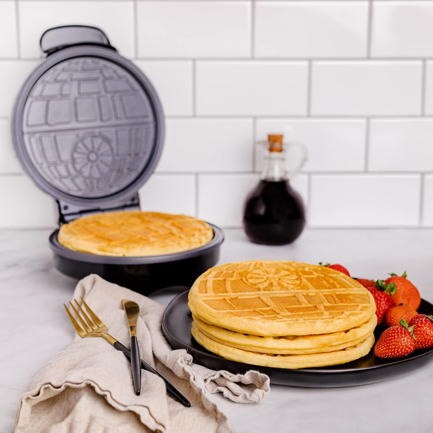 A waffle maker with a design resembling a sci-fi space station alongside waffles featuring the same pattern, accompanied by strawberries and syrup in the background.