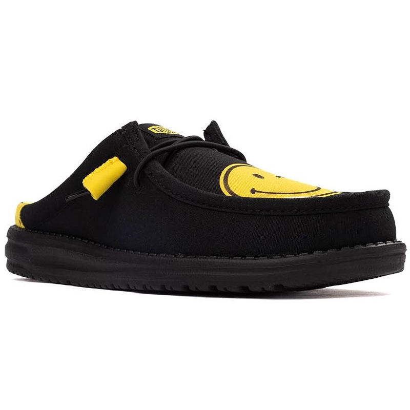 Hey Dude x SmileyWorld Mules feature a black design with a yellow smiley face on the upper and yellow accents on the heel and lace areas, offering a casual and cheerful look.