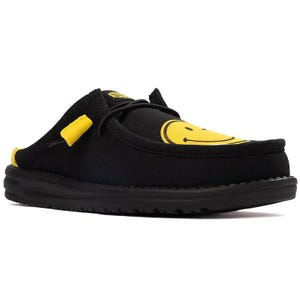 Hey Dude x SmileyWorld Mules feature a black design with a yellow smiley face on the upper and yellow accents on the heel and lace areas, offering a casual and cheerful look.