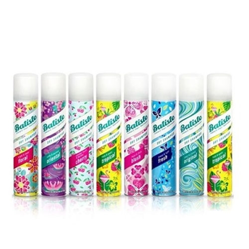 Seven cans of Batiste dry shampoo are lined up, featuring various scents such as floral, tropical, fresh, and blush, each with unique colorful designs on the cans.