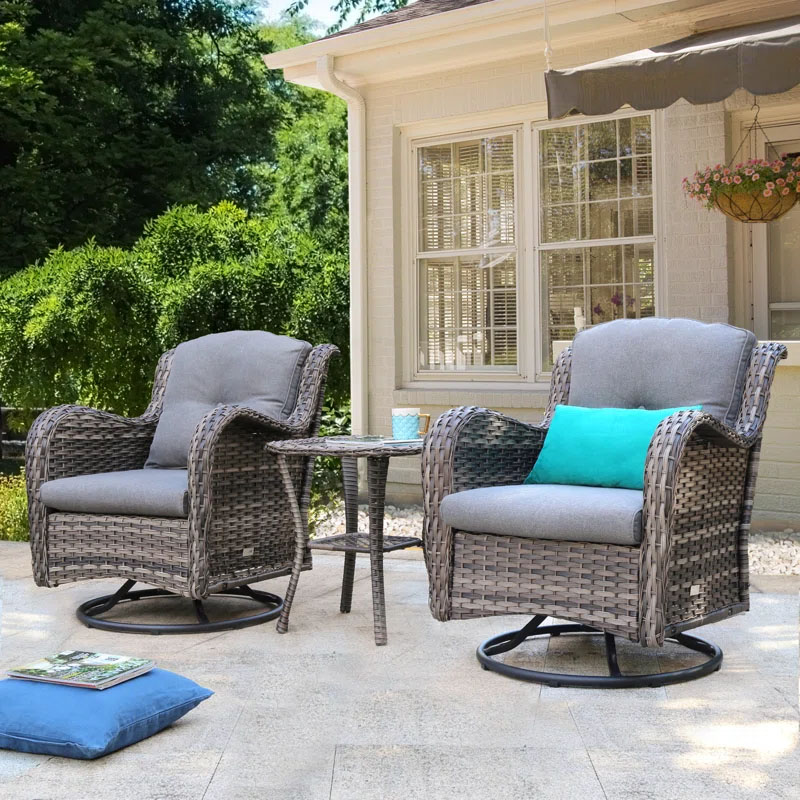 The 3pc Cushioned Rocker Set features two gray wicker swivel rockers with gray cushions, accompanied by a small round wicker side table. A blue throw pillow is placed on one of the chairs, all set on a light stone patio.