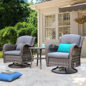 The 3pc Cushioned Rocker Set features two gray wicker swivel rockers with gray cushions, accompanied by a small round wicker side table. A blue throw pillow is placed on one of the chairs, all set on a light stone patio.
