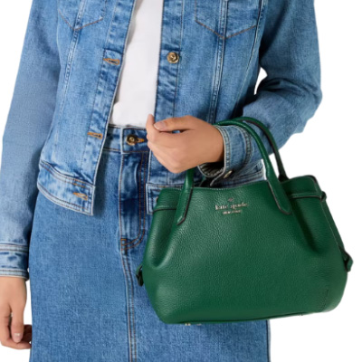 A green Kate Spade Small Dumpling Satchel with dual handles and the brand's logo on the front. The bag is shown held by a person wearing a denim jacket.