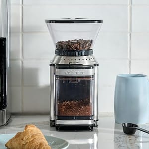 Coffee grinder with beans in the hopper and ground coffee in the container, next to a blue mug and a croissant on a marble countertop.