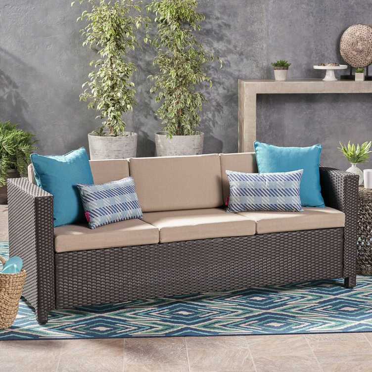 Brown wicker patio sofa with beige cushions, featuring two blue-bordered, geometric-patterned pillows and two solid blue square pillows. Placed on a blue and white patterned rug, with decorative plants and minimalist gray background.