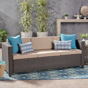 Brown wicker patio sofa with beige cushions, featuring two blue-bordered, geometric-patterned pillows and two solid blue square pillows. Placed on a blue and white patterned rug, with decorative plants and minimalist gray background.
