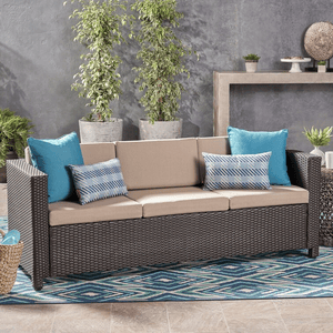 Brown wicker patio sofa with beige cushions, featuring two blue-bordered, geometric-patterned pillows and two solid blue square pillows. Placed on a blue and white patterned rug, with decorative plants and minimalist gray background.