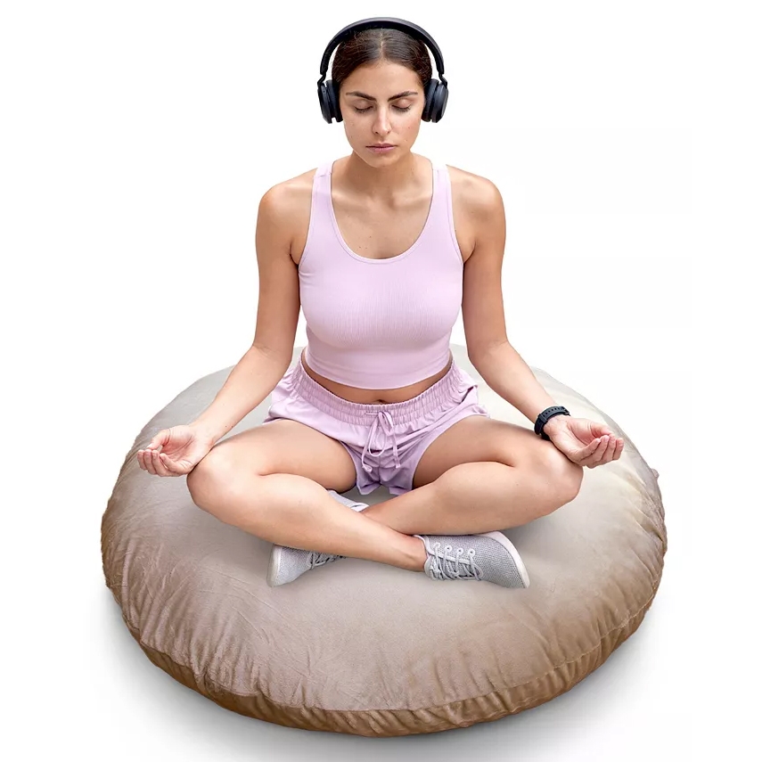 A person is meditating on a large round floor cushion while wearing wireless headphones and a smartwatch. They are dressed in a light pink tank top and shorts, sitting cross-legged.
