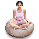 A person is meditating on a large round floor cushion while wearing wireless headphones and a smartwatch. They are dressed in a light pink tank top and shorts, sitting cross-legged.