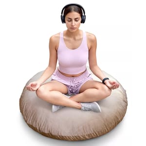 A person is meditating on a large round floor cushion while wearing wireless headphones and a smartwatch. They are dressed in a light pink tank top and shorts, sitting cross-legged.