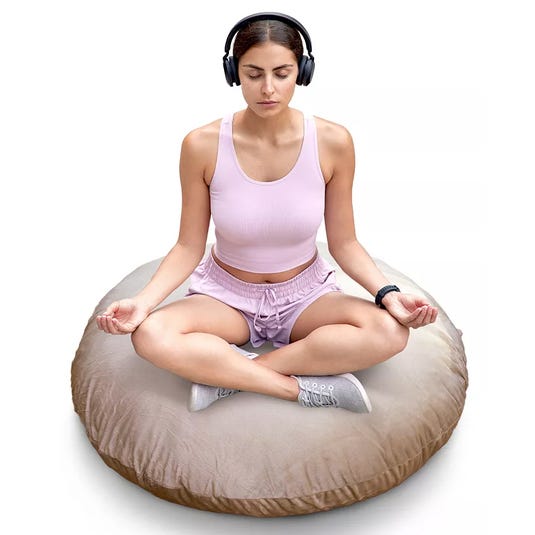 A person is meditating on a large round floor cushion while wearing wireless headphones and a smartwatch. They are dressed in a light pink tank top and shorts, sitting cross-legged.