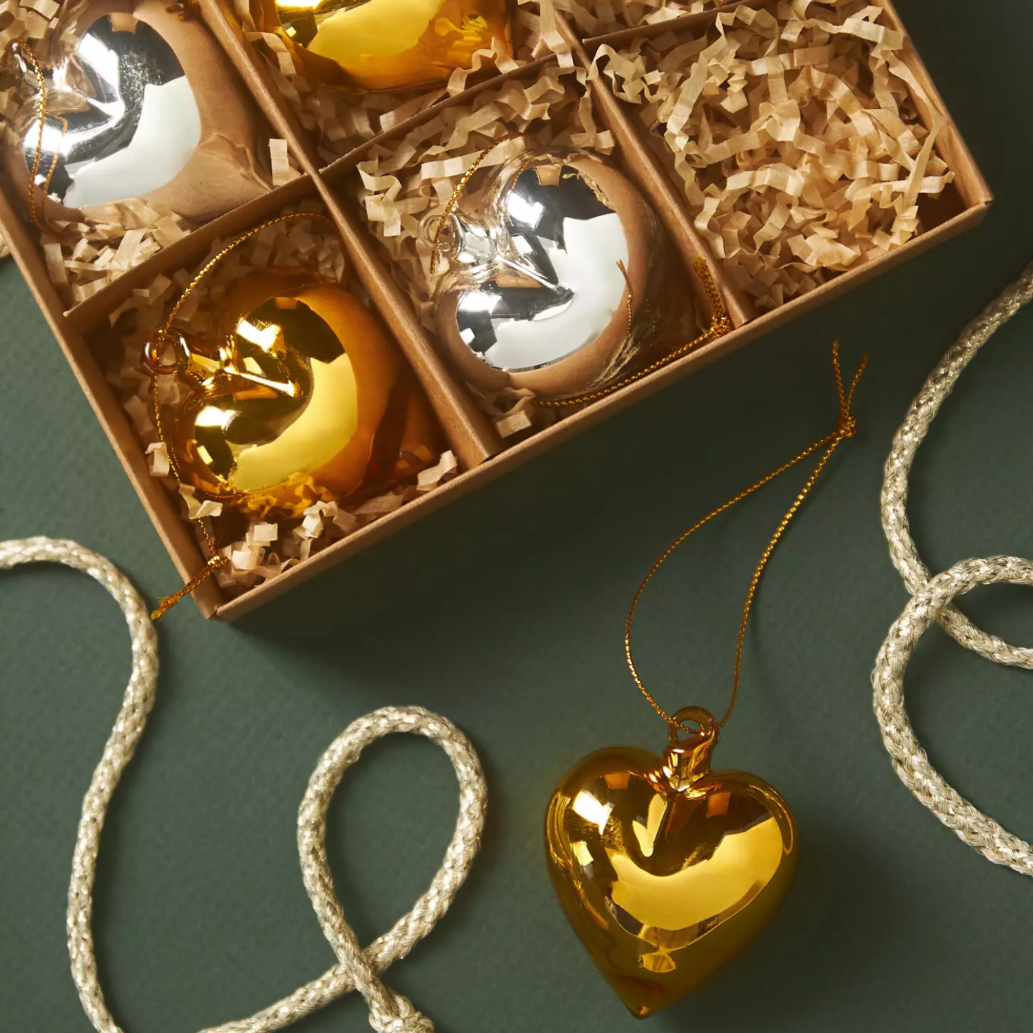 A set of six heart-shaped ornaments, three gold and three silver, packaged in a divided box with shredded paper padding. Each ornament has a string for hanging.