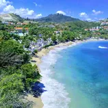 A scenic tropical beach with turquoise water, surrounded by lush greenery and hills with buildings in the background.
