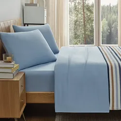Blue bed sheets and pillowcases with a striped bedspread, placed in a wooden bed frame. There's a wooden nightstand with books and a candle, next to a window showing a forest view.