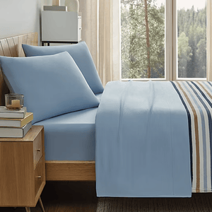 Blue bed sheets and pillowcases with a striped bedspread, placed in a wooden bed frame. There's a wooden nightstand with books and a candle, next to a window showing a forest view.