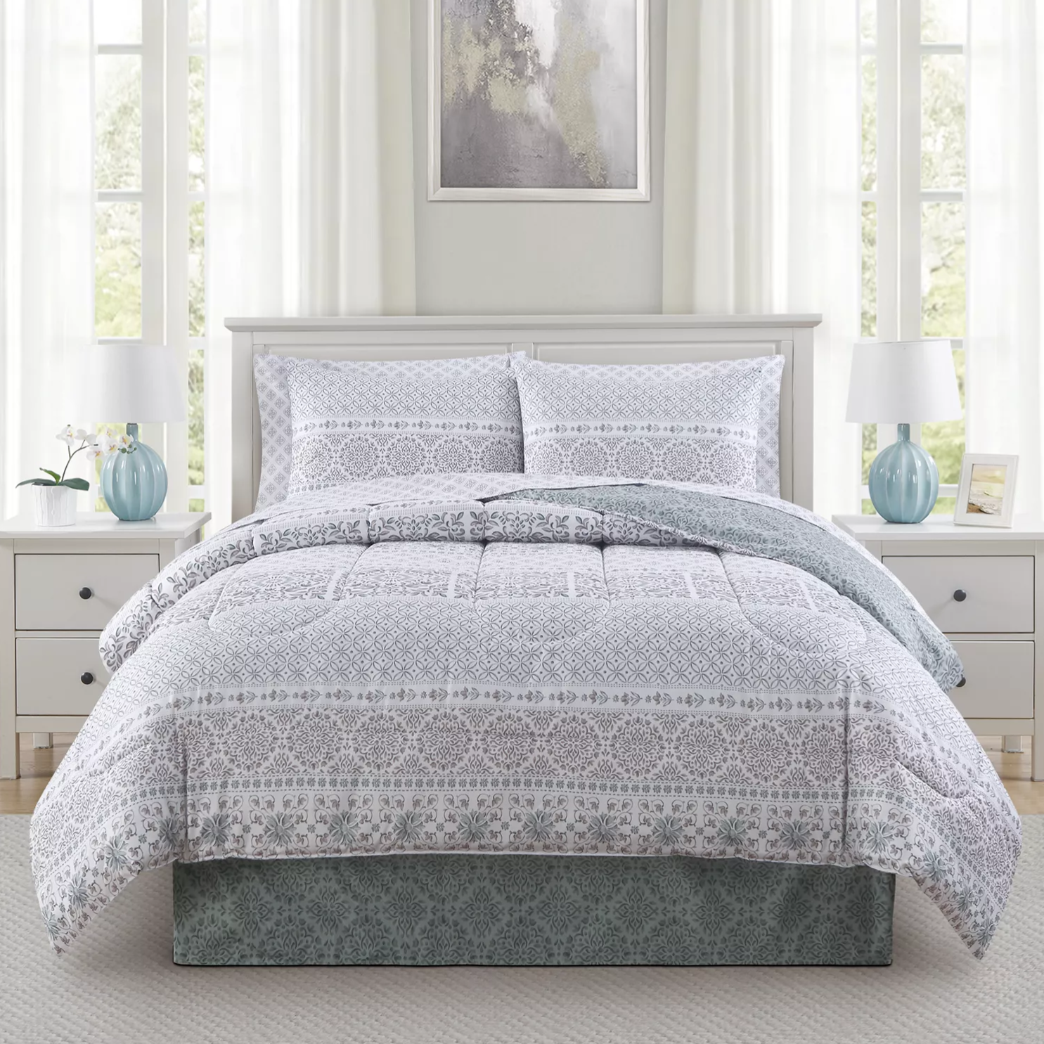 A bedroom features a white and gray patterned comforter set on a bed with matching pillow shams, flanked by two white nightstands with blue lamps. An abstract painting hangs above the bed, and soft white curtains cover windows, creating a light, airy ambiance.
