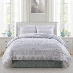 A bedroom features a white and gray patterned comforter set on a bed with matching pillow shams, flanked by two white nightstands with blue lamps. An abstract painting hangs above the bed, and soft white curtains cover windows, creating a light, airy ambiance.