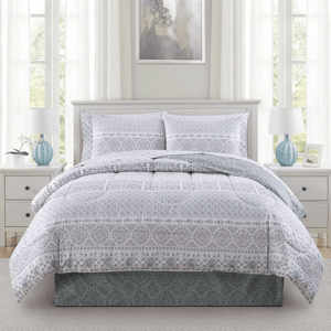 A bedroom features a white and gray patterned comforter set on a bed with matching pillow shams, flanked by two white nightstands with blue lamps. An abstract painting hangs above the bed, and soft white curtains cover windows, creating a light, airy ambiance.
