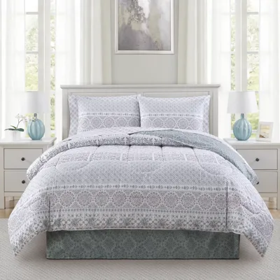 A bedroom features a white and gray patterned comforter set on a bed with matching pillow shams, flanked by two white nightstands with blue lamps. An abstract painting hangs above the bed, and soft white curtains cover windows, creating a light, airy ambiance.