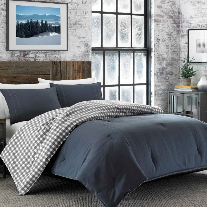 A cozy bedroom setup features a dark gray comforter with matching pillow shams, a checkered gray and white blanket, and a rustic wooden headboard. The room includes frosted windows, a small potted plant, and framed wall art.
