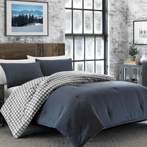 A cozy bedroom setup features a dark gray comforter with matching pillow shams, a checkered gray and white blanket, and a rustic wooden headboard. The room includes frosted windows, a small potted plant, and framed wall art.