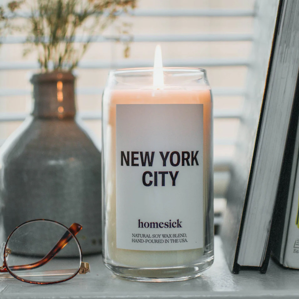 A lit candle labeled \“NEW YORK CITY\“ by Homesick is on display, surrounded by a book, eyeglasses, and a vase with flowers.