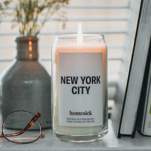 A lit candle labeled \“NEW YORK CITY\“ by Homesick is on display, surrounded by a book, eyeglasses, and a vase with flowers.