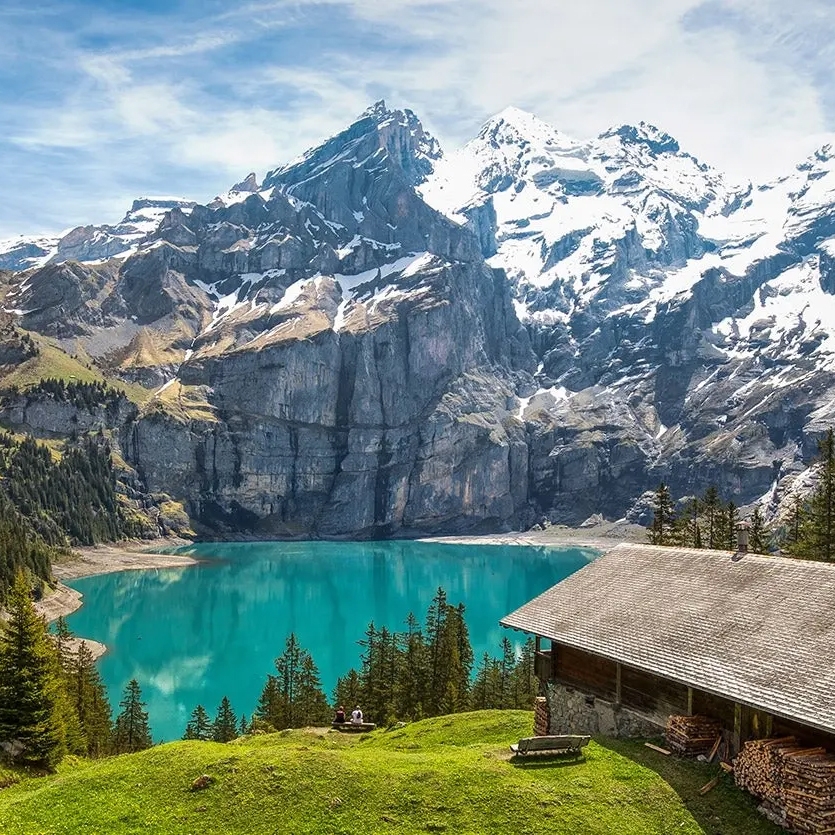 A pristine mountain lake with a backdrop of snow-capped peaks and a wooden cabin surrounded by greenery.