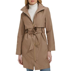 A woman is wearing a light brown trench coat with a front zipper and tie belt, paired with a white turtleneck sweater and blue jeans.