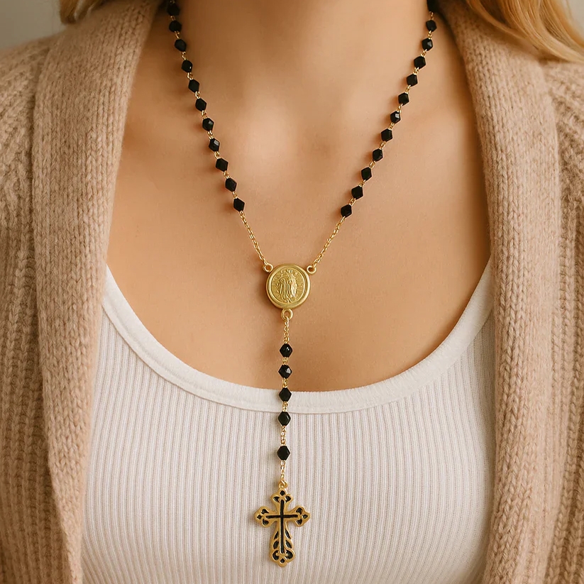 A gold-tone rosary necklace with black beads and a cross pendant, featuring a medallion at the center.