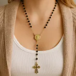 A gold-tone rosary necklace with black beads and a cross pendant, featuring a medallion at the center.