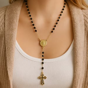 A gold-tone rosary necklace with black beads and a cross pendant, featuring a medallion at the center.