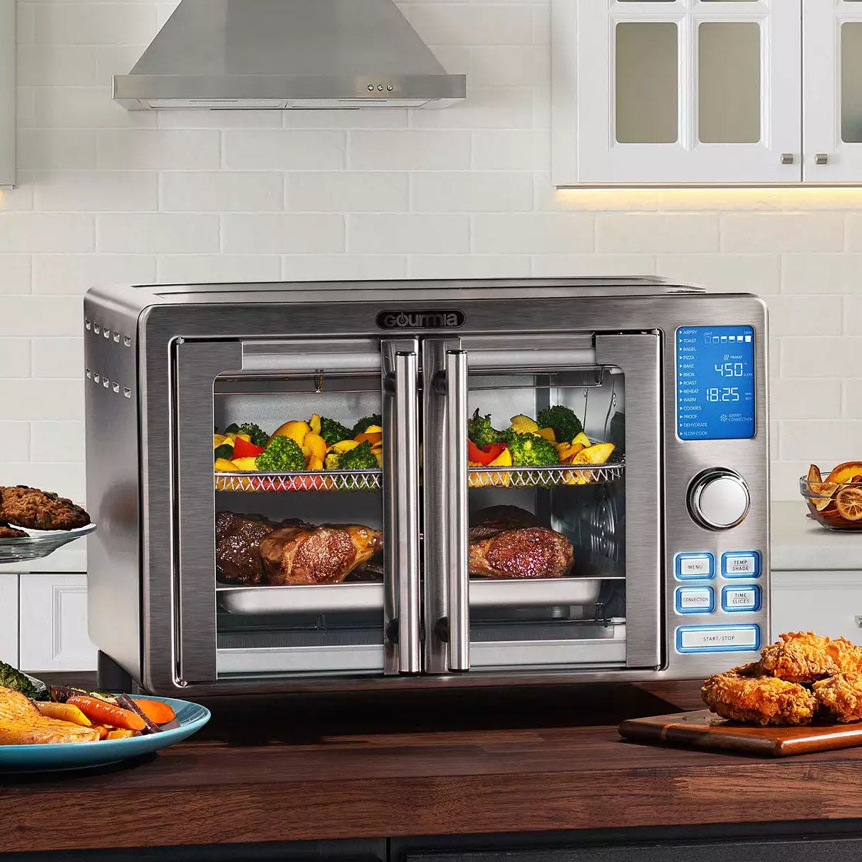 A stainless steel Gourmia air fryer oven with a digital display is shown cooking steaks and vegetables. It features multiple buttons and French doors, positioned on a kitchen countertop with fried chicken and more food around it.
