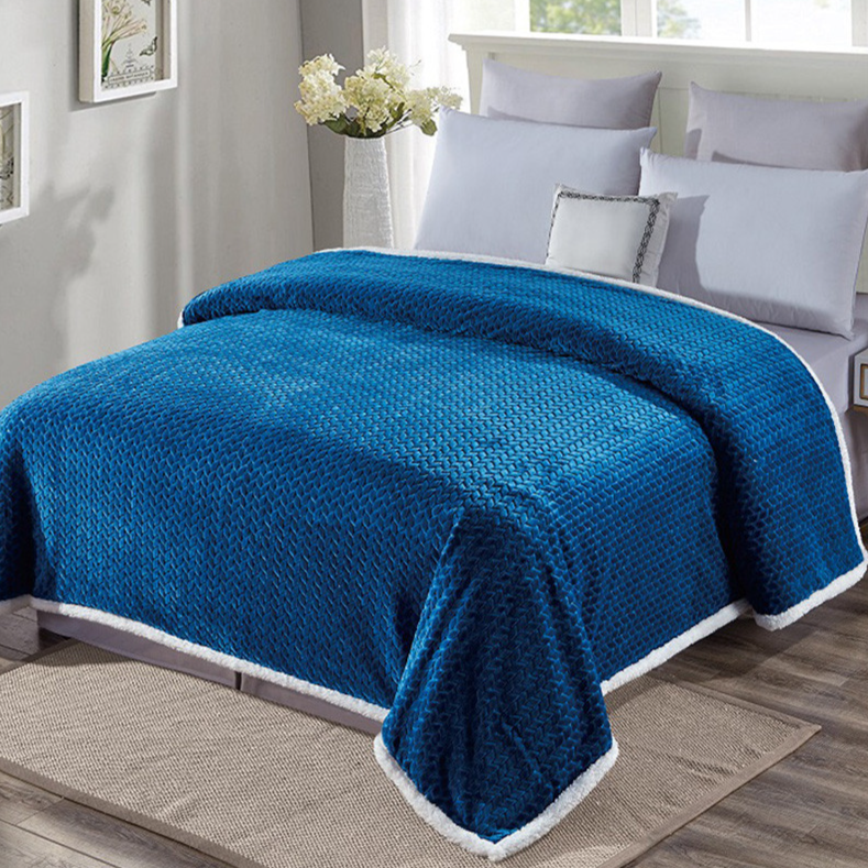 A blue plush blanket with a textured pattern and white trim covers a bed with white pillows in a sunlit bedroom. White flowers in a vase and small framed artworks decorate the room.
