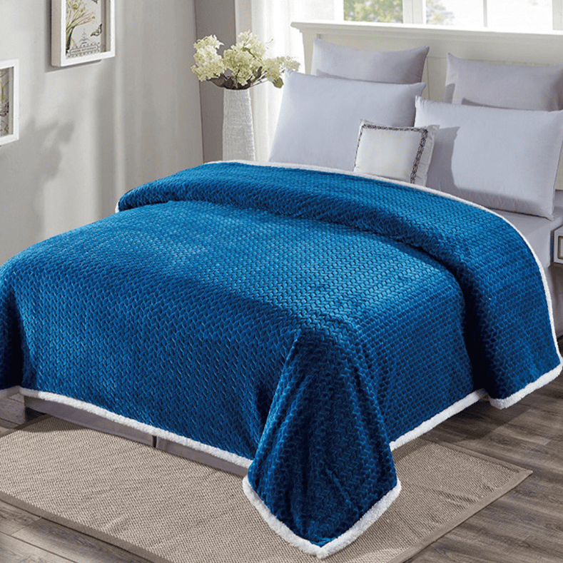 A blue plush blanket with a textured pattern and white trim covers a bed with white pillows in a sunlit bedroom. White flowers in a vase and small framed artworks decorate the room.