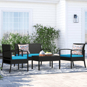 The 4-Piece Carlyssa Rattan Patio Set includes a loveseat, two armchairs, and a coffee table, all with black rattan frames. The set features blue cushions and decorative patterned pillows, set on an outdoor rug and surrounded by greenery.
