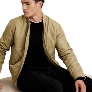 A man wears a light brown bomber jacket from Banana Republic Factory, featuring ribbed cuffs, a round collar, and front pockets, paired with a black shirt and pants.
