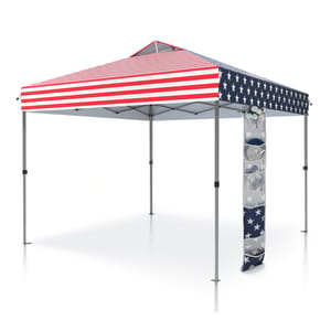A pop-up canopy tent with an American flag design features red and white stripes on the roof and blue sides with white stars. A storage accessory with transparent pockets is attached to one leg.