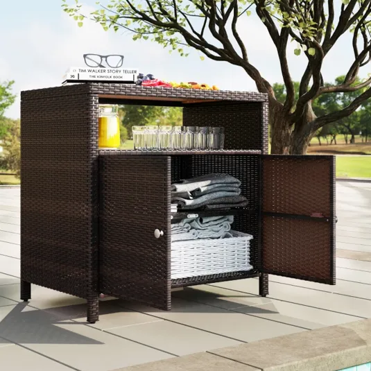 Poolside wicker cabinet with open shelving displays folded towels, glasses, books, and a pitcher of juice under a tree.