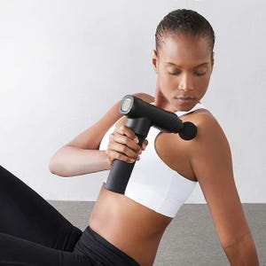 A woman is using a handheld percussion massage gun on her arm.