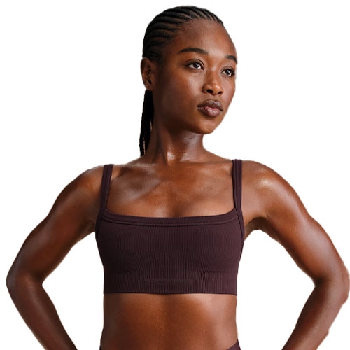 A woman is wearing a dark brown sports bra with thin straps and has sweat on her skin, suggesting athletic activity.