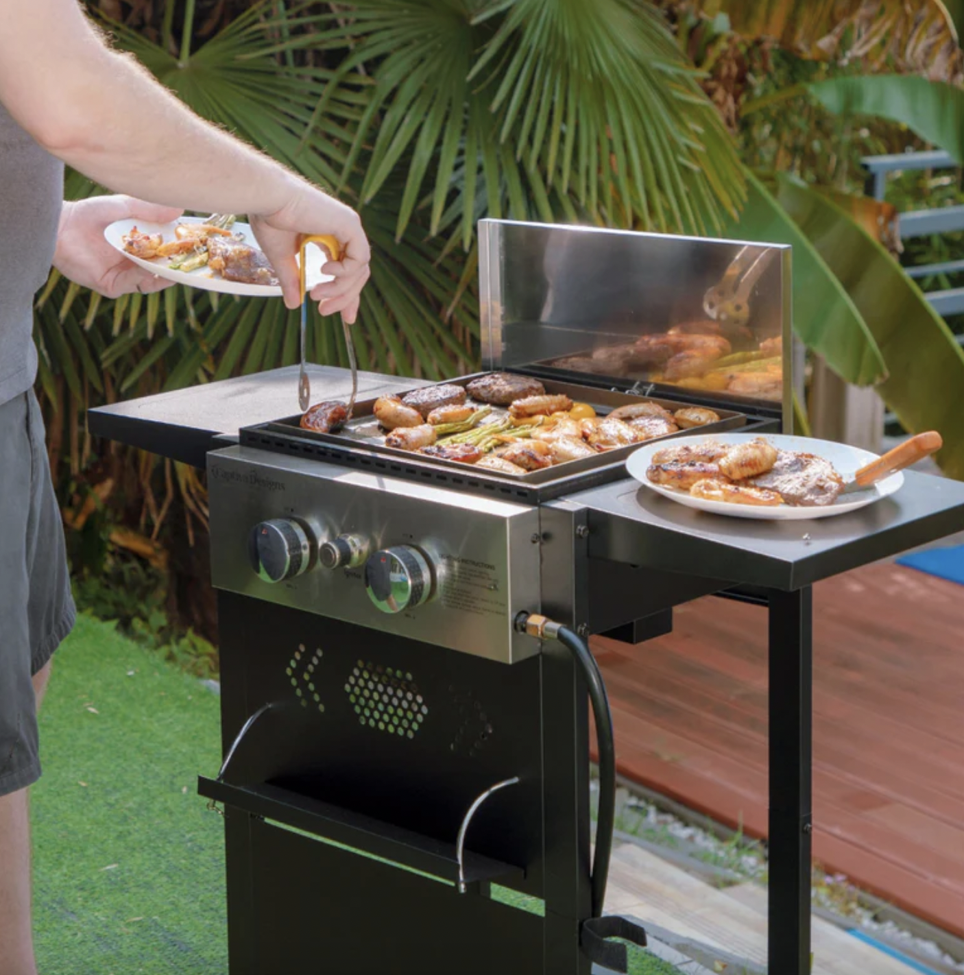 A two-burner gas griddle grill with a stainless steel control panel and gas hose connection, featuring a wind guard and side shelf. A person is grilling various meats and vegetables, with plates holding cooked items on the side shelf.