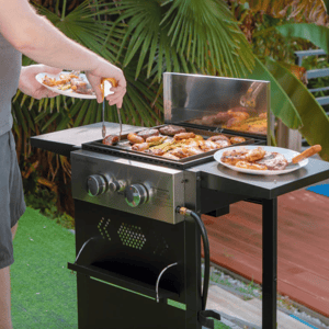 A two-burner gas griddle grill with a stainless steel control panel and gas hose connection, featuring a wind guard and side shelf. A person is grilling various meats and vegetables, with plates holding cooked items on the side shelf.