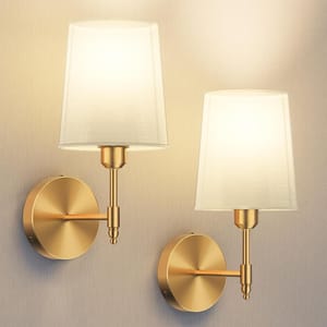 A pair of gold wall sconces with white fabric drum shades, mounted on round backplates, providing soft ambient lighting.